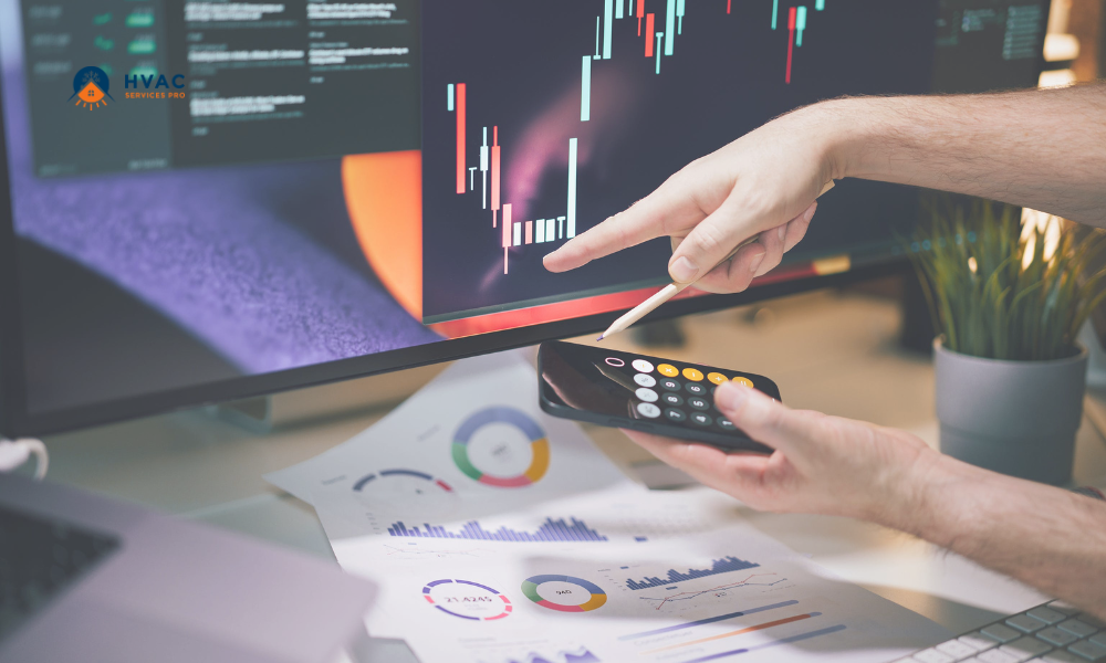 Person analyzing stock charts on a computer monitor, pointing with a pen. Smartphone calculator and financial graphs on a desk, suggesting financial analysis.