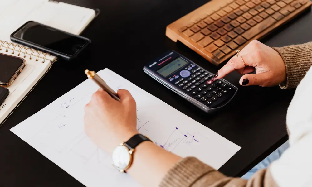 A person wearing a brown sweater uses a calculator and writes on paper at a desk, with a keyboard, phone, and notebook nearby, conveying focus and calculation.